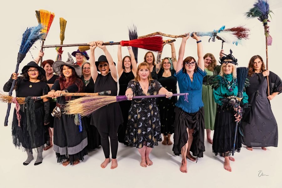 Members of the Forest City Dance Circle holding their decorated brooms overhead in a horizontal line, with two more brooms held out in front at waist height, in black and jewel-toned costume.