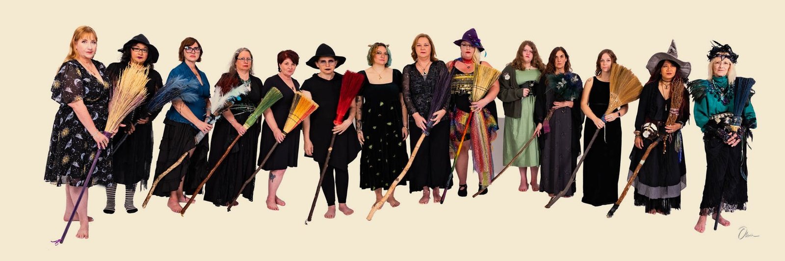 The Forest City Dance Circle standing in a long line in black and jewel-toned seasonal costume, most of them holding a handmade broom.