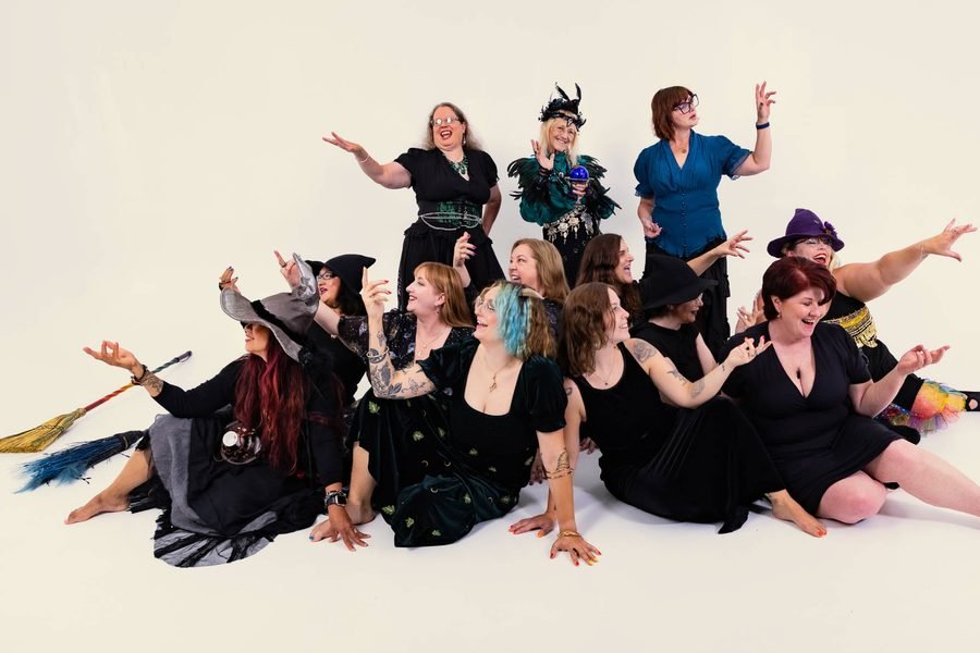 Dancers of the Forest City Dance Circle clustered together, some seated on the floor and three standing behind, hands raised in a dance gesture, laughing.