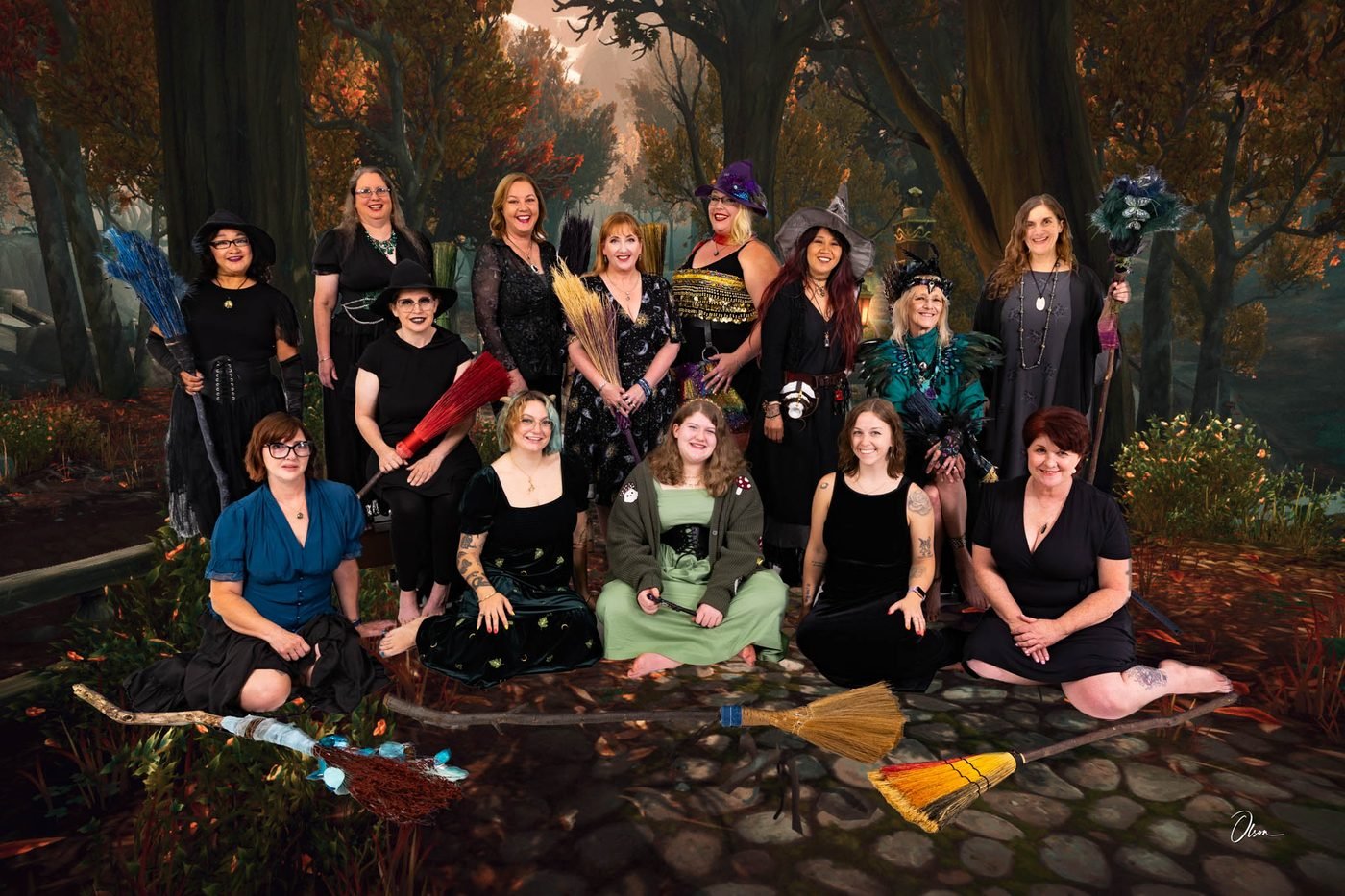 The Forest City Dance Circle gathered on a cobbled woodland path beneath autumn trees, in black and jewel-toned costume, their handmade brooms laid out in front of them.